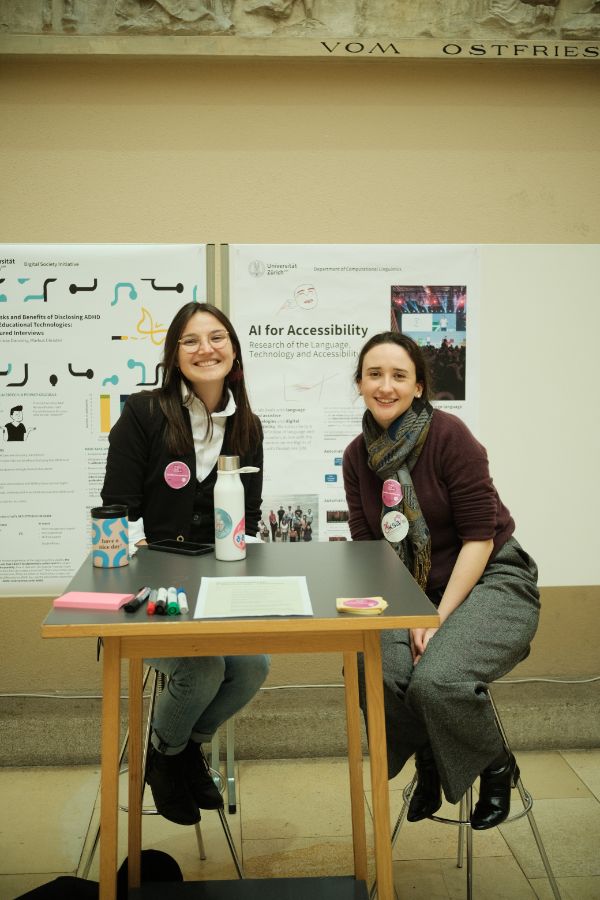 Infostand Accessibility-Forschung: Zwei Personen sitzen lächelnd an einem kleinen Infotisch vor einem Poster mit der Aufschrift „AI for Accessibility“ im Lichthof der UZH.